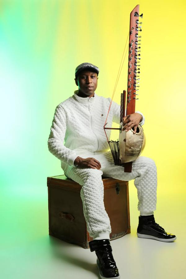 A person sits on a storage box wearing a white quilted tracksuit and holding a kora