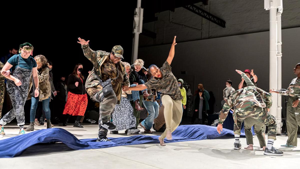 A group of dancers dressed in camouflage clothing are pictured mid motion. A long undulating strip of blue cloth stretches across the middle of the floor. 