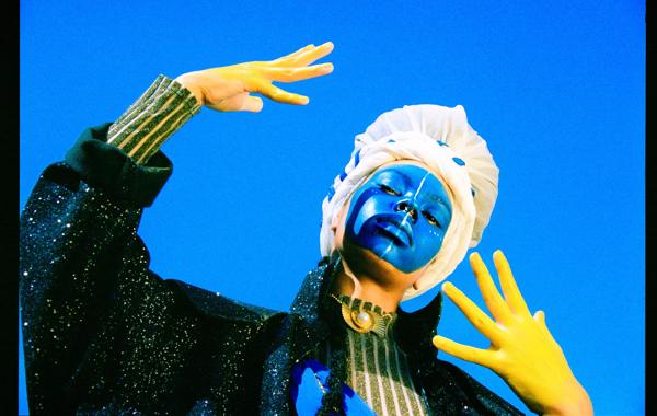 A model, their face painted blue and hands painted yellow, poses against a blue sky