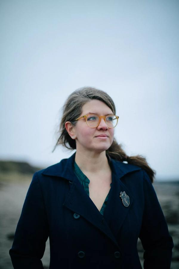 Author Jessie Greengrass stands on a beach  in a blue jacket with her hair pulled back and glasses on.