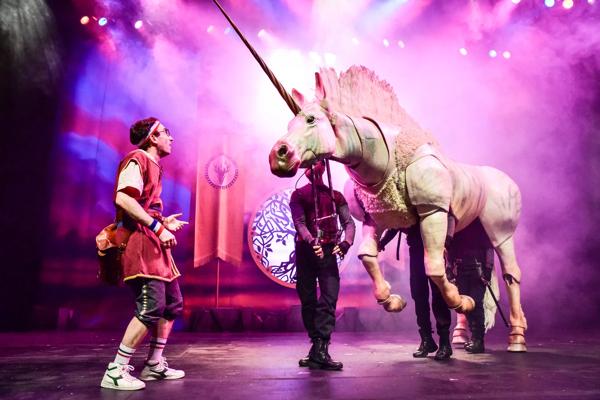 A man stands on stage alongside a puppet unicorn held by three other people 
