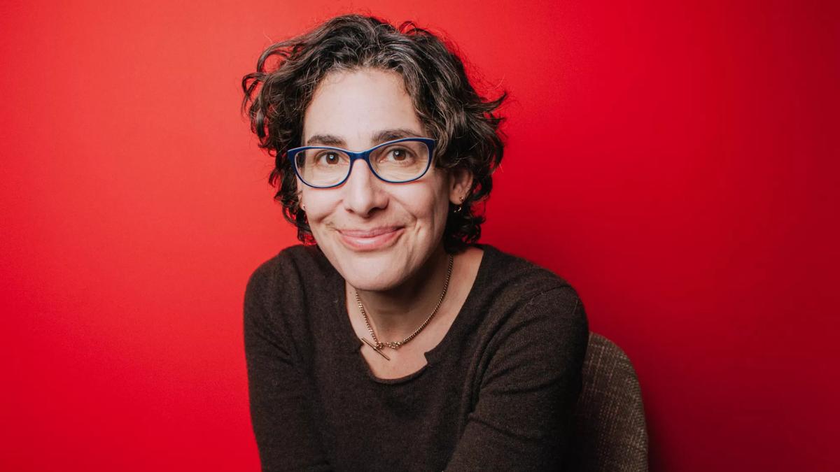 Sarah Koenig sits in front of a red background wearing a grey top