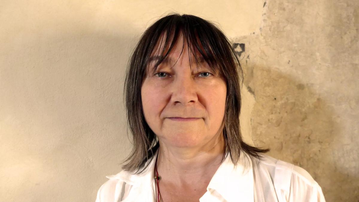 Portrait of author Ali Smith. She is in a white collared shirt in front of a textured beige wall. 
