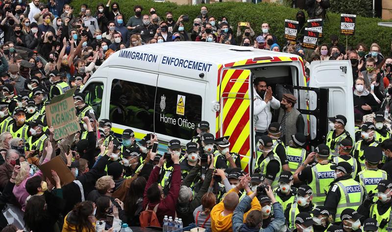 Two men are released from the back of an immigration enforcement van. The van is surrounded by police officers, and in turn the police officers are surrounded by hundreds of people. Many of the people are applauding, one of the men leaving the van is raising his hands in thanks