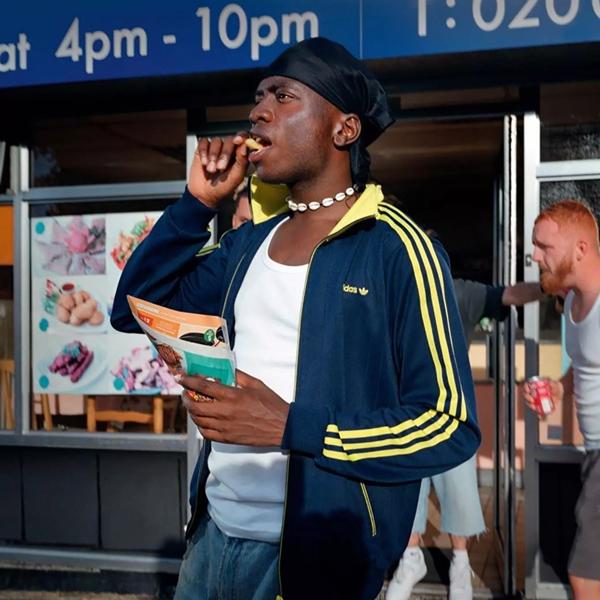 Musician Master Peace standing outside of a shop holding a paper cone of chips and putting one chip into his mouth. He wears a navy blue jacket with yellow stripes and a black do-rag on his head