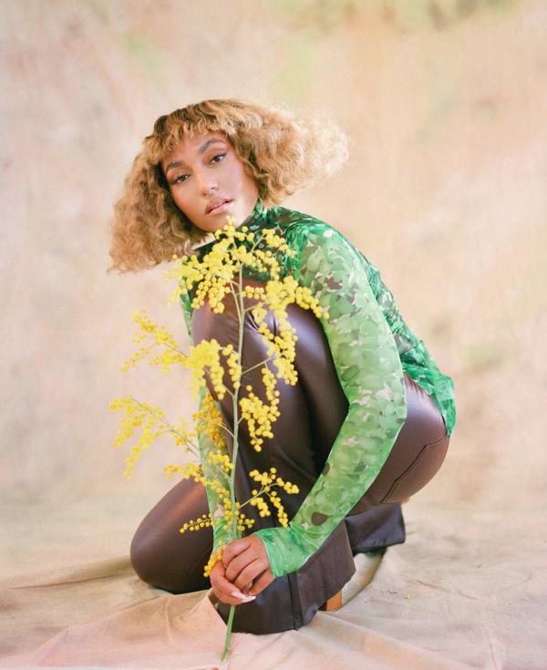 A person with blond wavy hair poses for a portrait wearing a green long-sleeve shirt and holding a yellow flower
