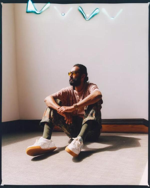 A man with a beard and yellow tinted sunglasses sits on the floor with his elbows resting on his knees looking away from camera. 