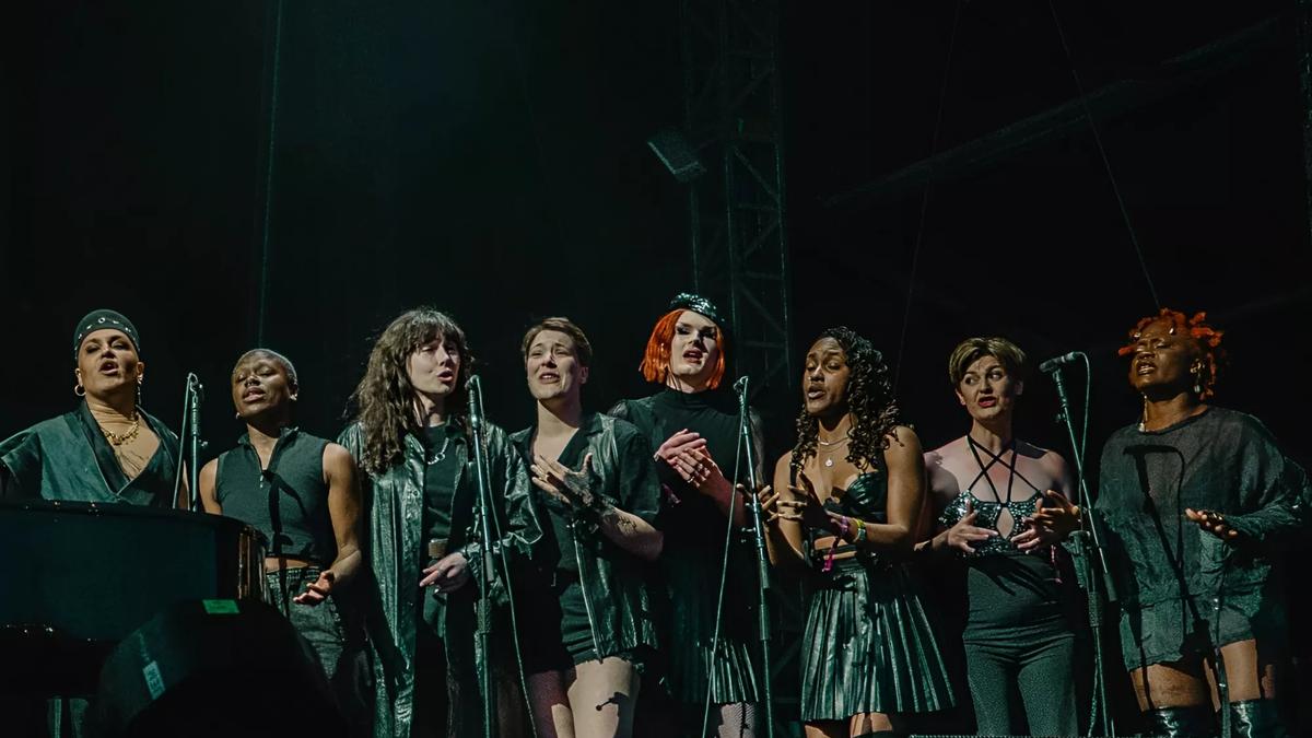 The Trans Voices choir all stand in front of microphones wearing black singing. 