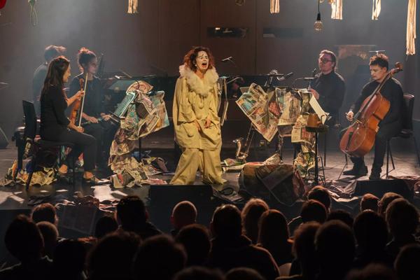 Patricia Kopatchinskaja is dressed as Pierrot the clown on an expressionistic set with a group of musicians playing strings and woodwind
