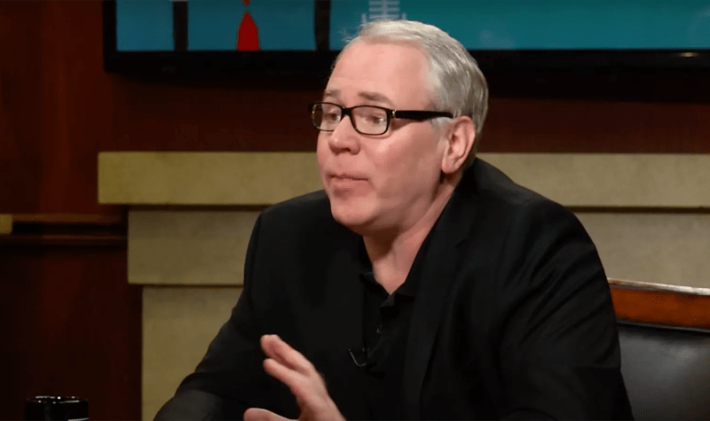 Author Brett Easton Ellis wears a black shirt and suit as he is interviewed in a TV studio next to a wooden desk