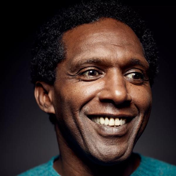 Lemn Sissay smiling and wearing a blue jumper against a black background.