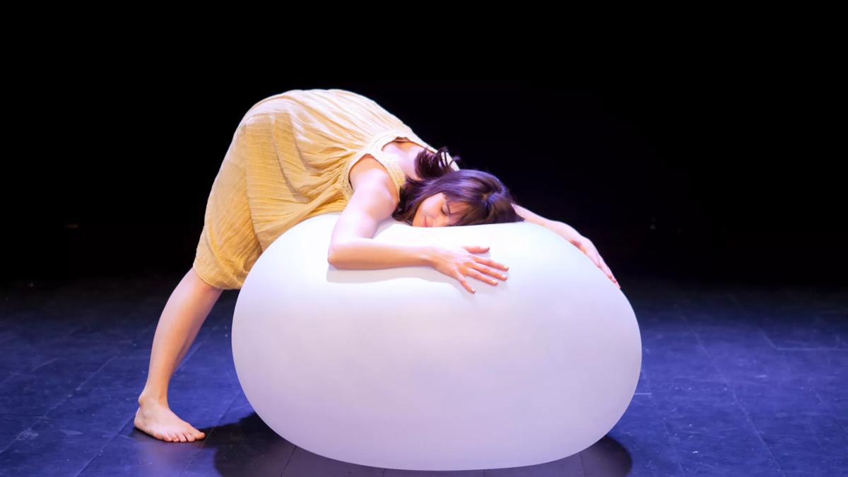 Female performer in a yellow dress leans her head on a large white balloon.