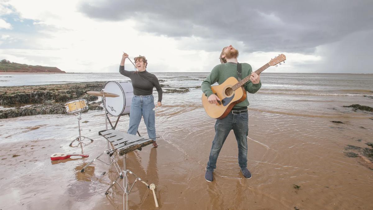 A man and a woman surrounded by instruments throw their heads back shouting on the beach. The man is on the right of the image playing an acoustic guitar and the woman is on the left of the image playing percussion. 