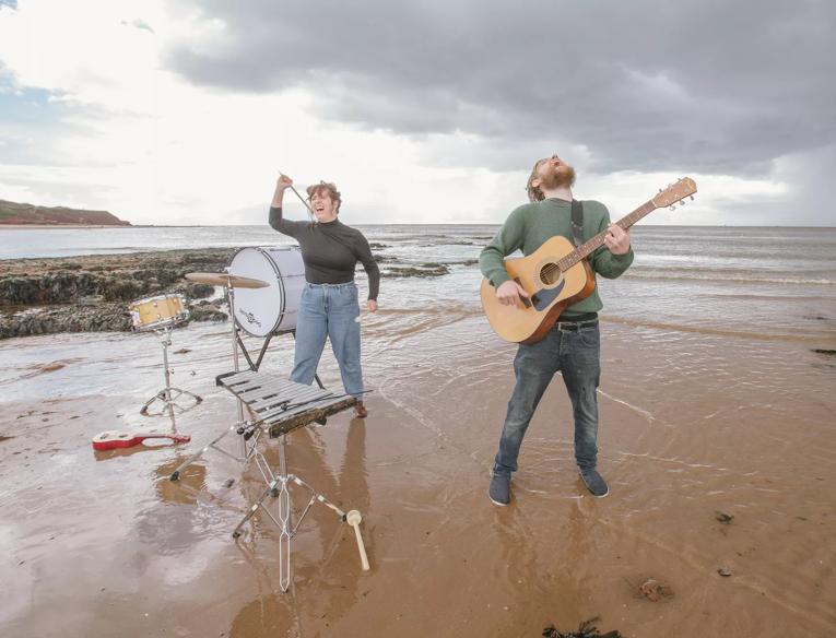 A man and a woman surrounded by instruments throw their heads back shouting on the beach. The man is on the right of the image playing an acoustic guitar and the woman is on the left of the image playing percussion. 
