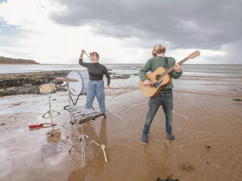 A man and a woman surrounded by instruments throw their heads back shouting on the beach. The man is on the right of the image playing an acoustic guitar and the woman is on the left of the image playing percussion. 