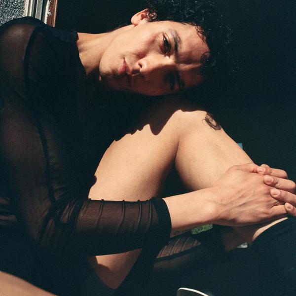 A man with his head leaning on his knee, wearing a sheer black top