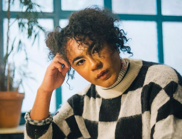 Travis Alabanza leans on a table in a black and white chequered jumper in front of a window
