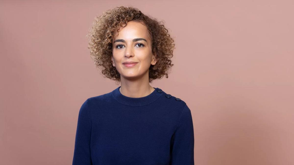 Leila has cropped, curly honey coloured hair and smiles to camera. She wears a blue jumper and is standing against a pink/brown background. 