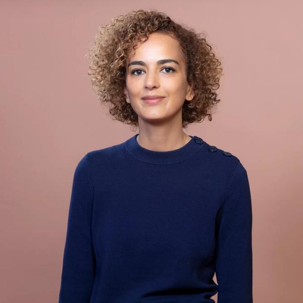 Leila has cropped, curly honey coloured hair and smiles to camera. She wears a blue jumper and is standing against a pink/brown background. 