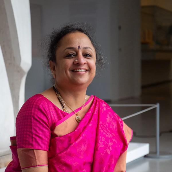 Supriya Nagarajan wearing a pink sari sitting down with a microphone and smiling.