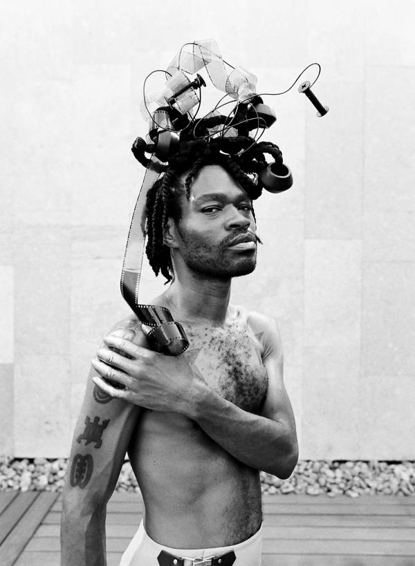 A topless Black man with an elaborate headress holds his right shoulder with his left arm 