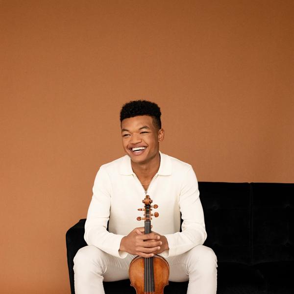 Randall Goosby in white holding a violin and sitting on a black sofa