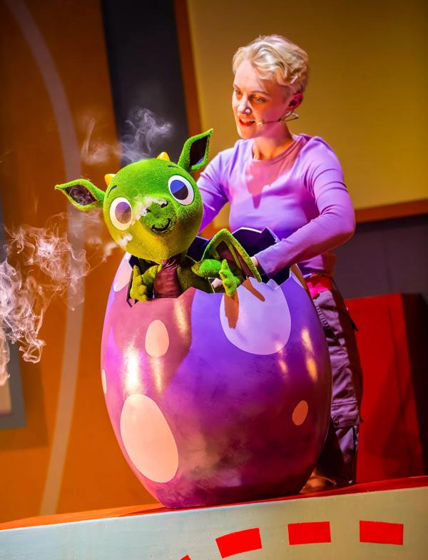 Actor holds a green dragon puppet which is hatching out of a purple egg, with smoke coming out of its nose.