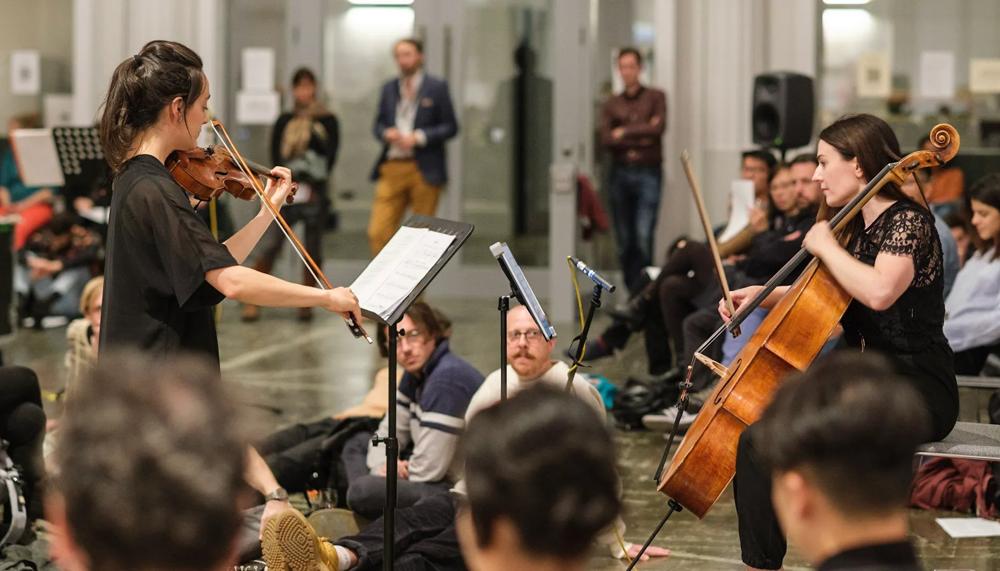 Two women: one plays the violin and one the cello. They are in what looks like a rehearsal room or a gym. Many people sat on chairs and on the floor are intent on watching them play.