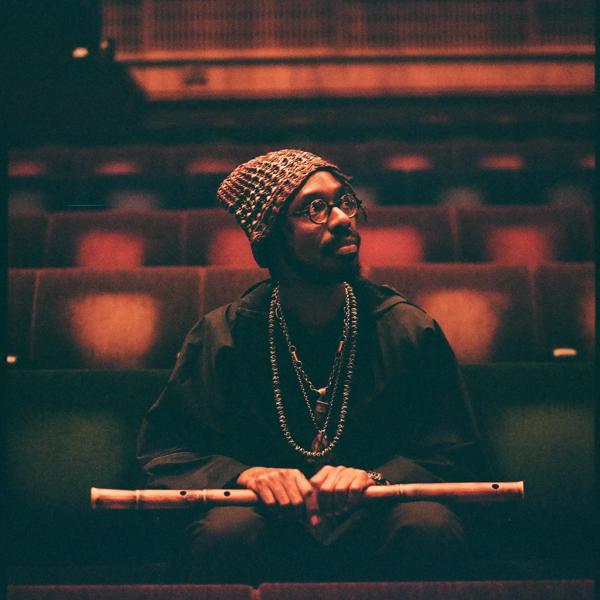 A man wearing a beanie and glasses looking away from the camera holding a wooden flute