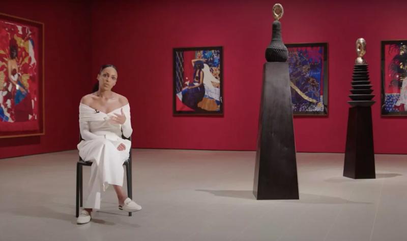Artist Lina Iris Viktor wearing a long white dress sits among her own works within Hayward Gallery's exhibition, In the Black Fantastic. The walls are painted red and there are paintings hung on them, whilst there are two sculptural pieces to the artist's left.