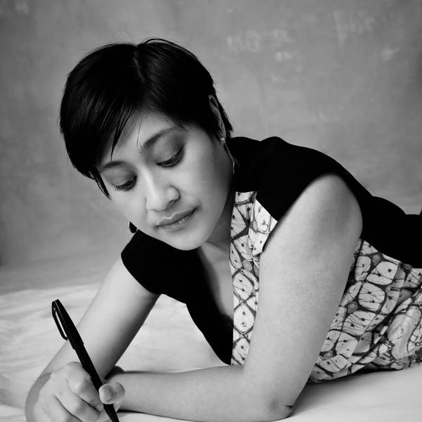 Khairani Barokka, depicted in black and white, lays across a white surface and holds a pen wit which she has written on the same surface