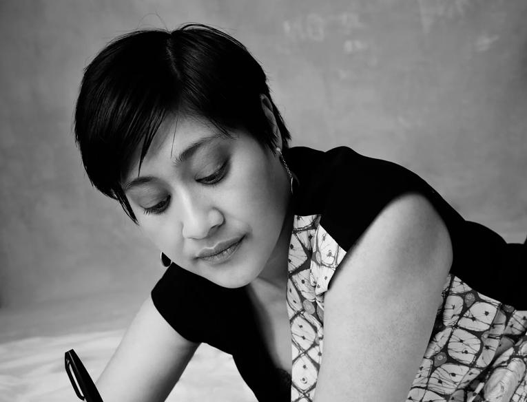 Khairani Barokka, depicted in black and white, lays across a white surface and holds a pen wit which she has written on the same surface