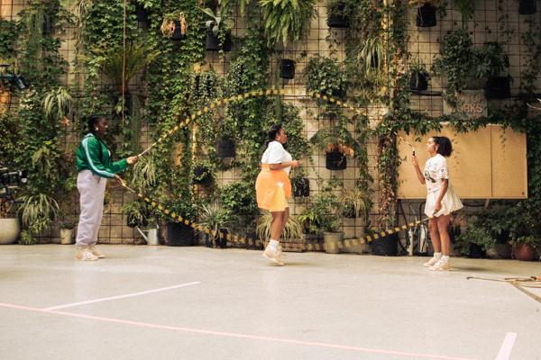three black girls are playing with two skipping ropes with a back drop of lots of plants on a wall 