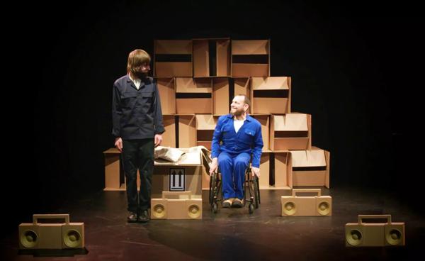 Person wearing a blue boiler suit sitting in a wheelchair looking at someone standing next to them. There are several cardboard boomboxes around them.