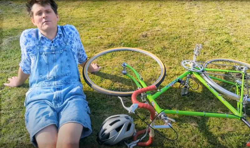 The musician David Gibb, a White man in his 30s wearing light blue shirt and shorts sits on the grass next to his bike, a green racer with red handlebars