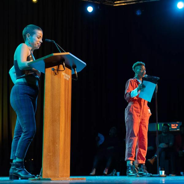 The person on the left is standing behind a podium and the one on the right is speaking to a microphone with a paper in hand. Other emerging artist panelist sitting at the back o the stage 
