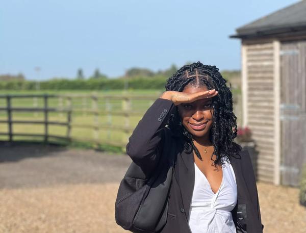A Black woman in a black blazer and white t shirt holds her hand shielding her eyes from the sun and looks to camera. 