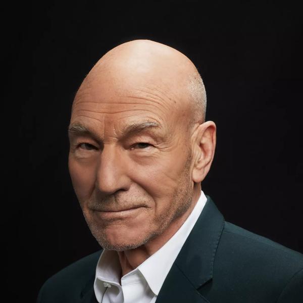 Patrick Stewart wears a sharp, dark suit over a white shirt and is pictured smiling to camera at a three quarter angle. He is bald with a faint stubble and blue eyes. 