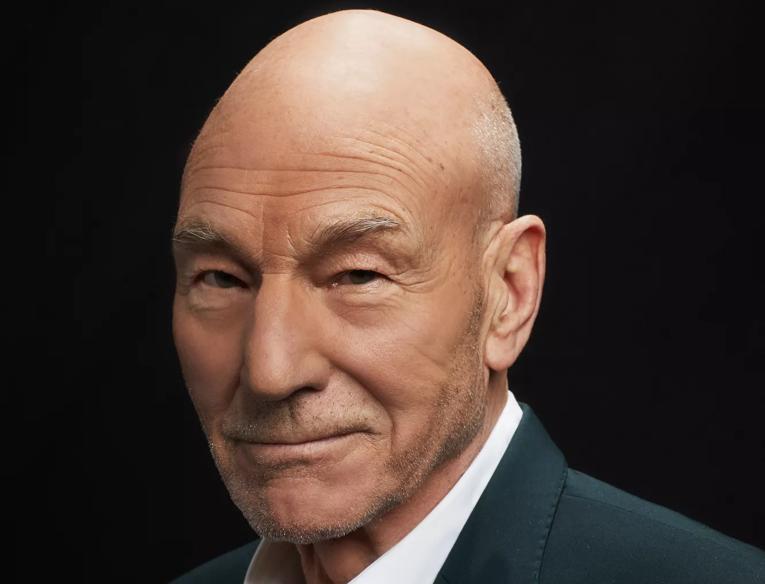 Patrick Stewart wears a sharp, dark suit over a white shirt and is pictured smiling to camera at a three quarter angle. He is bald with a faint stubble and blue eyes. 