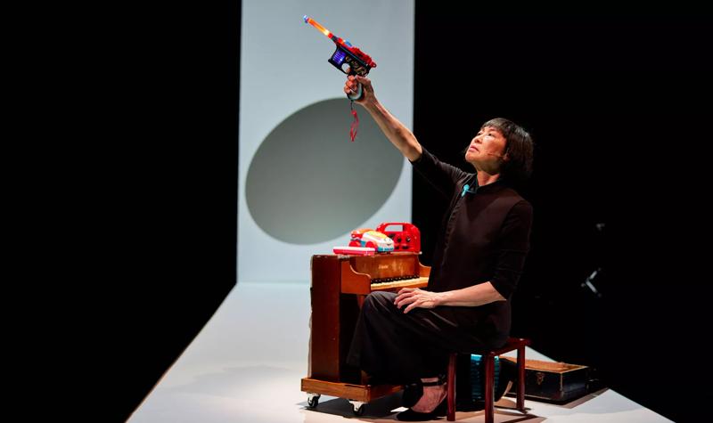woman sits at a child's size piano, she points a red and blue toy gun into the air.