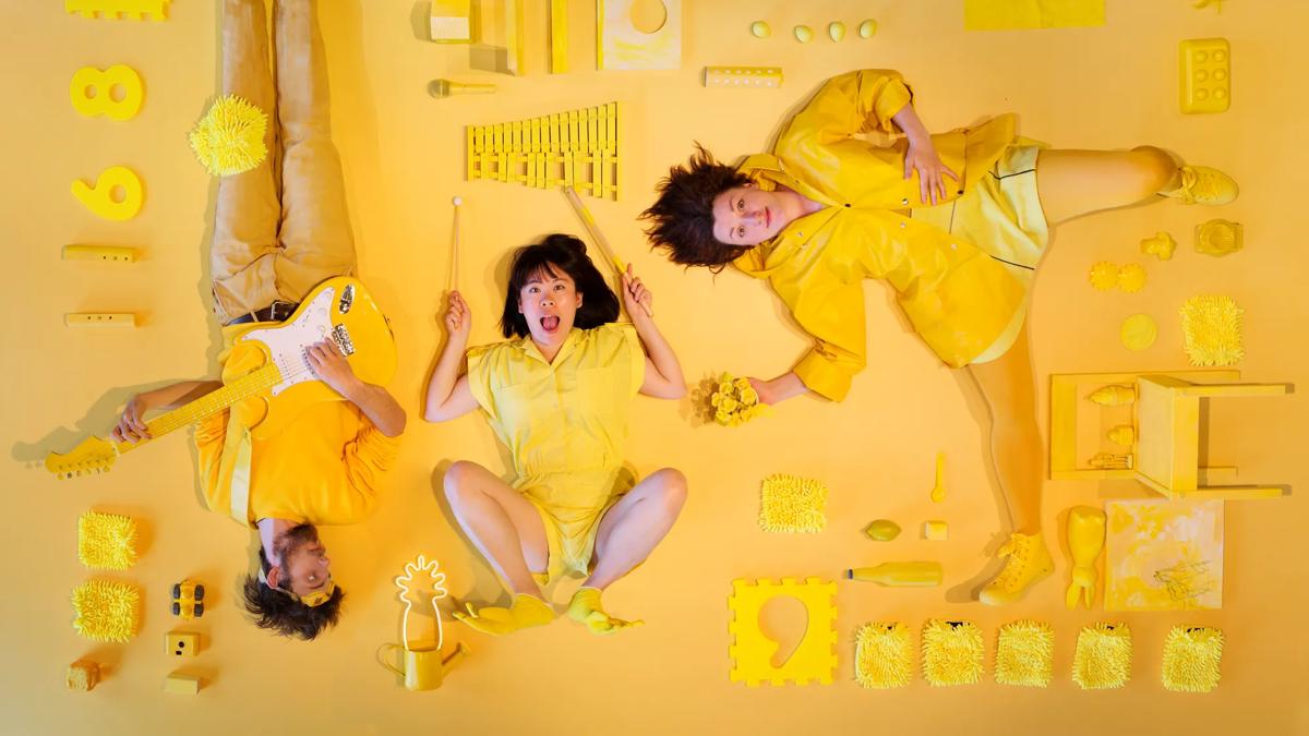 Three people lying on a yellow background surrounded by an assortment of yellow objects, wearing yellow clothes.