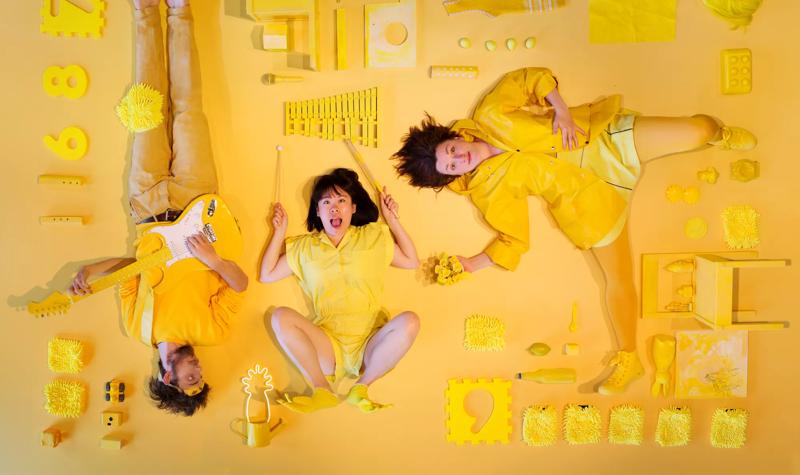 Three people lying on a yellow background surrounded by an assortment of yellow objects, wearing yellow clothes.