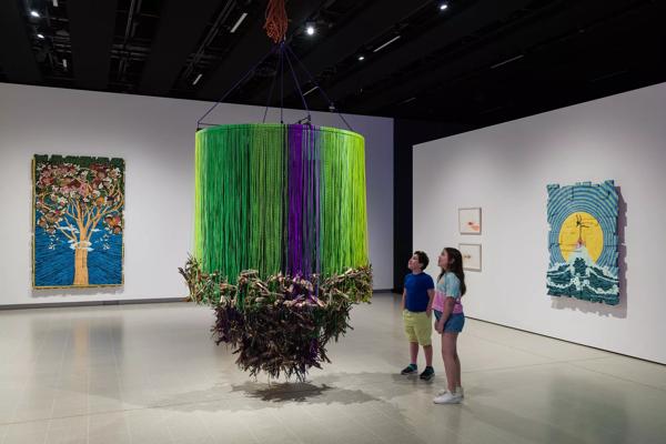 Two children looking at an art piece made up of green and purple shoe laces with wooden sticks hanging at the bottom