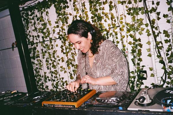 A DJ wearing a patterned shirt and smiling stands behind the decks in front of an ivy lined wall