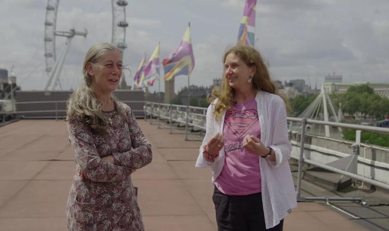 Activists Heather and Helen B, middle-aged White women, give an interview on the roof of the Royal Festival Hall. Their flag creation Union Jill and the London Eye can be seen behind them