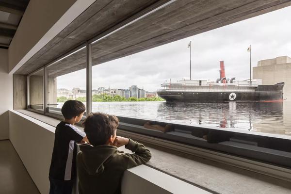 An installation view of kids watching a boat floating in black water with a brutalist building in the background.