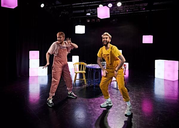 Two actors on stage looking excited. One wears yellow dungarees and the other wears pink dungarees. There are tables and chairs behind them.
