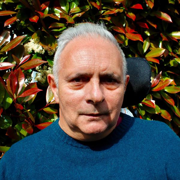 Hanif Kureishi wearing a blue jumper standing against a leafy background.