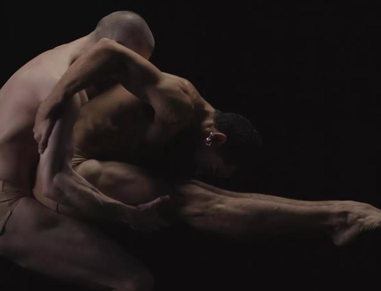 Two dancers, one in a full body bend with pointed toes, with the person behind them is supporting them by holding them under the legs. Both are wearing only nude coloured underwear.