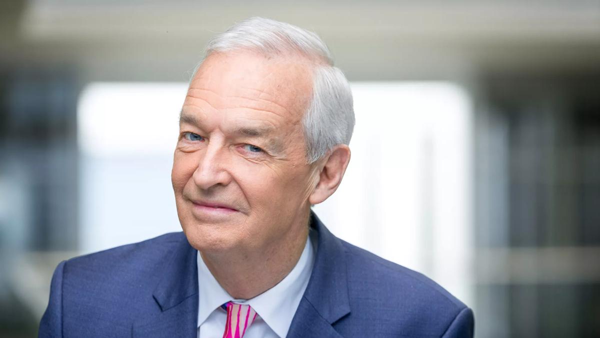 Broadcaster Jon Snow smiles to the camera. He is wearing a blue suit, a pink striped tie and the background of the image is blurred so only Jon Snow is in focus. 